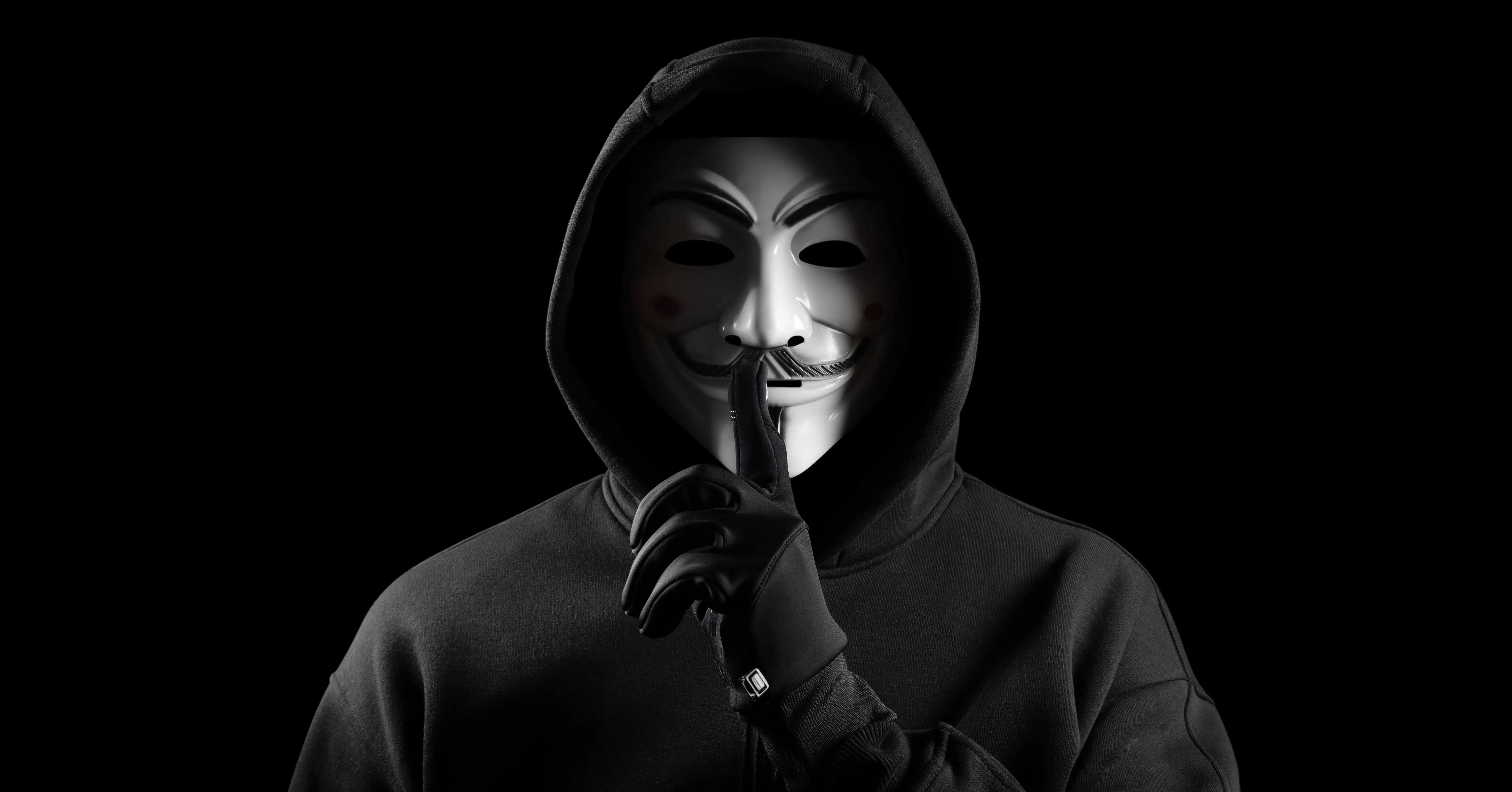 Person i mörk hoodie och handskar håller ett finger framför munnen i en tystnadsgest mot en svart bakgrund