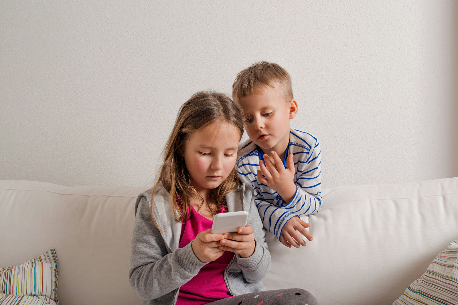 Ett flicka sitter i en soffa och tittar på en mobil. En pojke står bakom soffan och tittar på mobilen också.