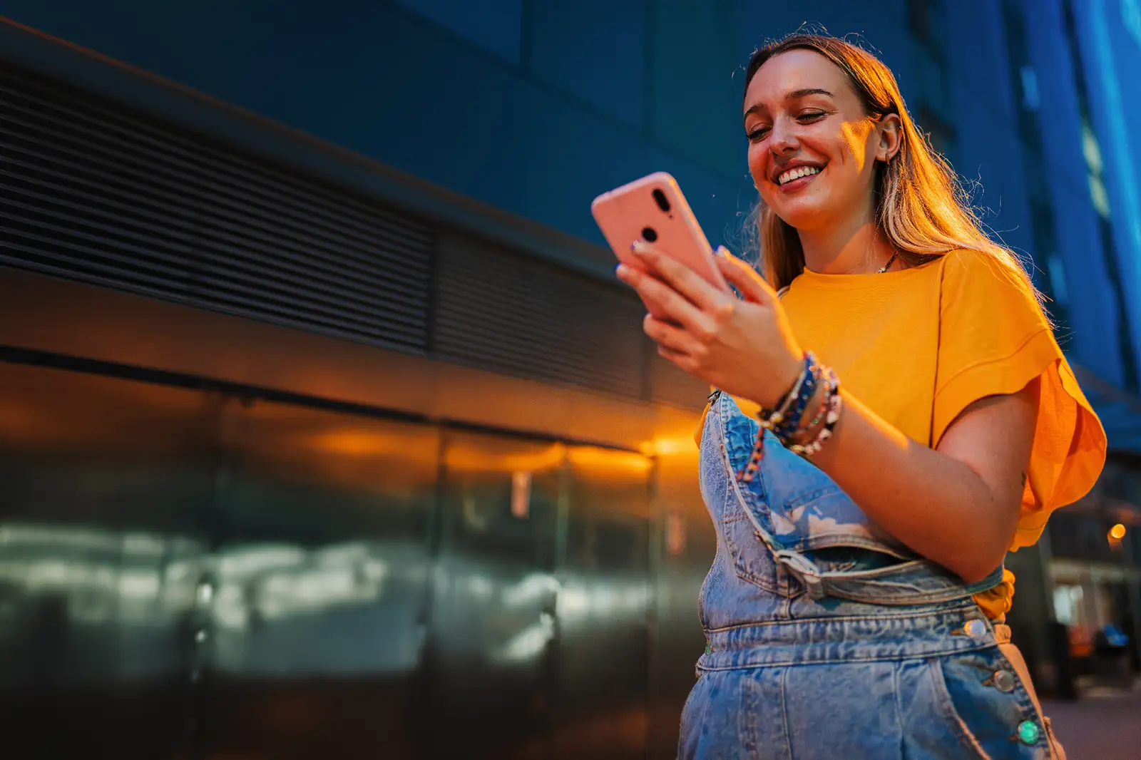 En person i gul topp står utomhus i stadsmiljö i skymningen och håller en mobiltelefon i handen.