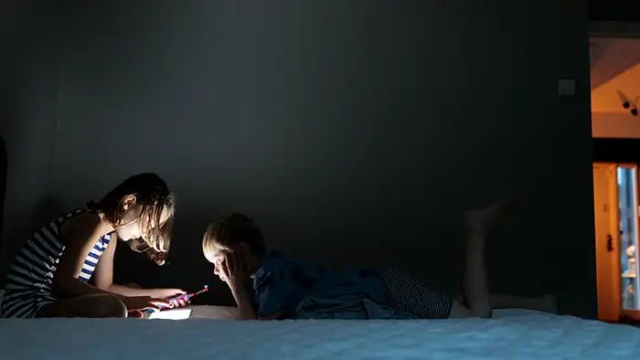 Två barn tittar på sina skärmar i ett mörkt rum och skärmarna lyser starkt